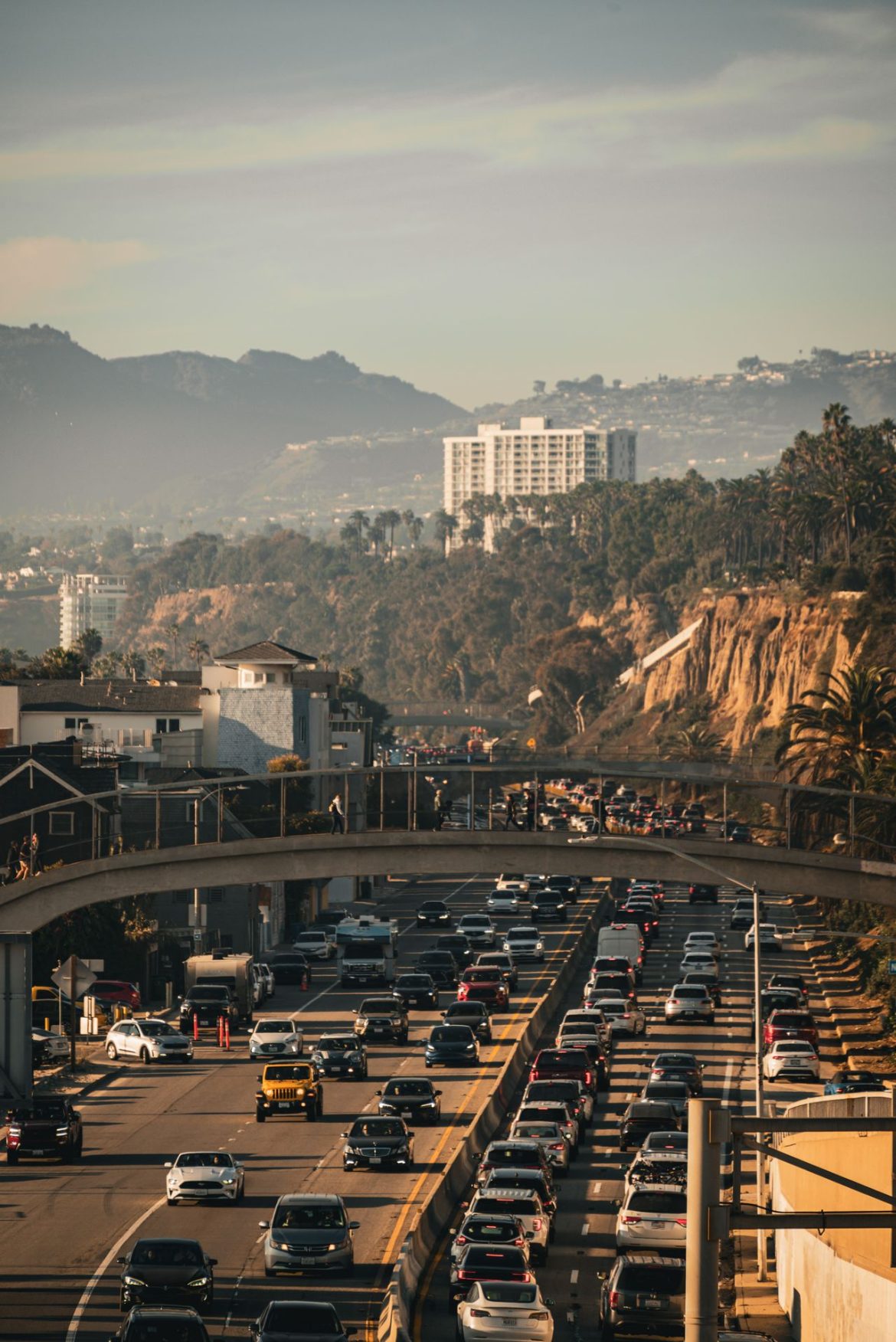 Rush-hour traffic on a coastal California highway with a footbridge overhead, palm-covered cliffs, and a hazy city skyline in the distance.