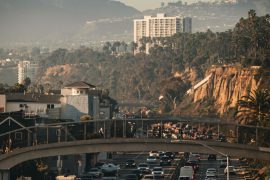 Rush-hour traffic on a coastal California highway with a footbridge overhead, palm-covered cliffs, and a hazy city skyline in the distance.